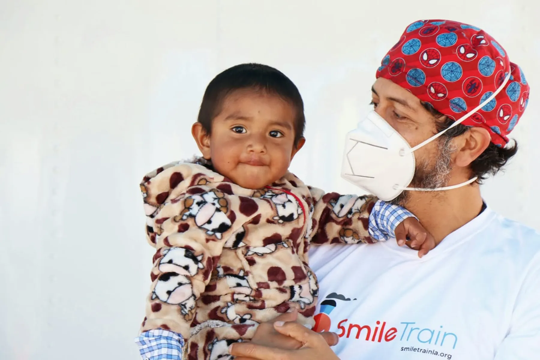 Dr. Nico Sierra con mascarilla y gorro quirúrgico sostiene en brazos a un niño pequeño que mira a cámara