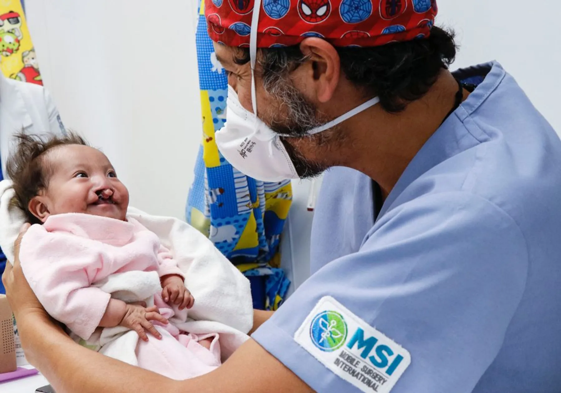 Dr. Nico Sierra con gorro quirúrgico sonríe a un bebé con fisura labial que le devuelve la sonrisa en consulta