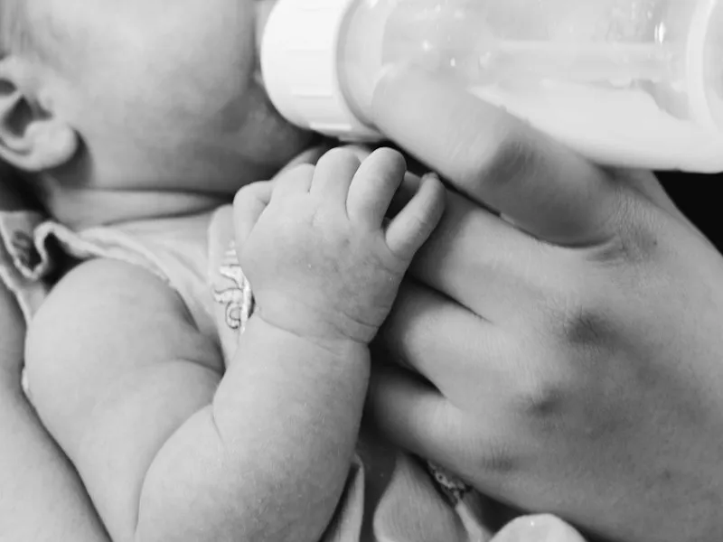 Manos de un adulto sosteniendo un biberón junto a las manos de un bebé, en blanco y negro